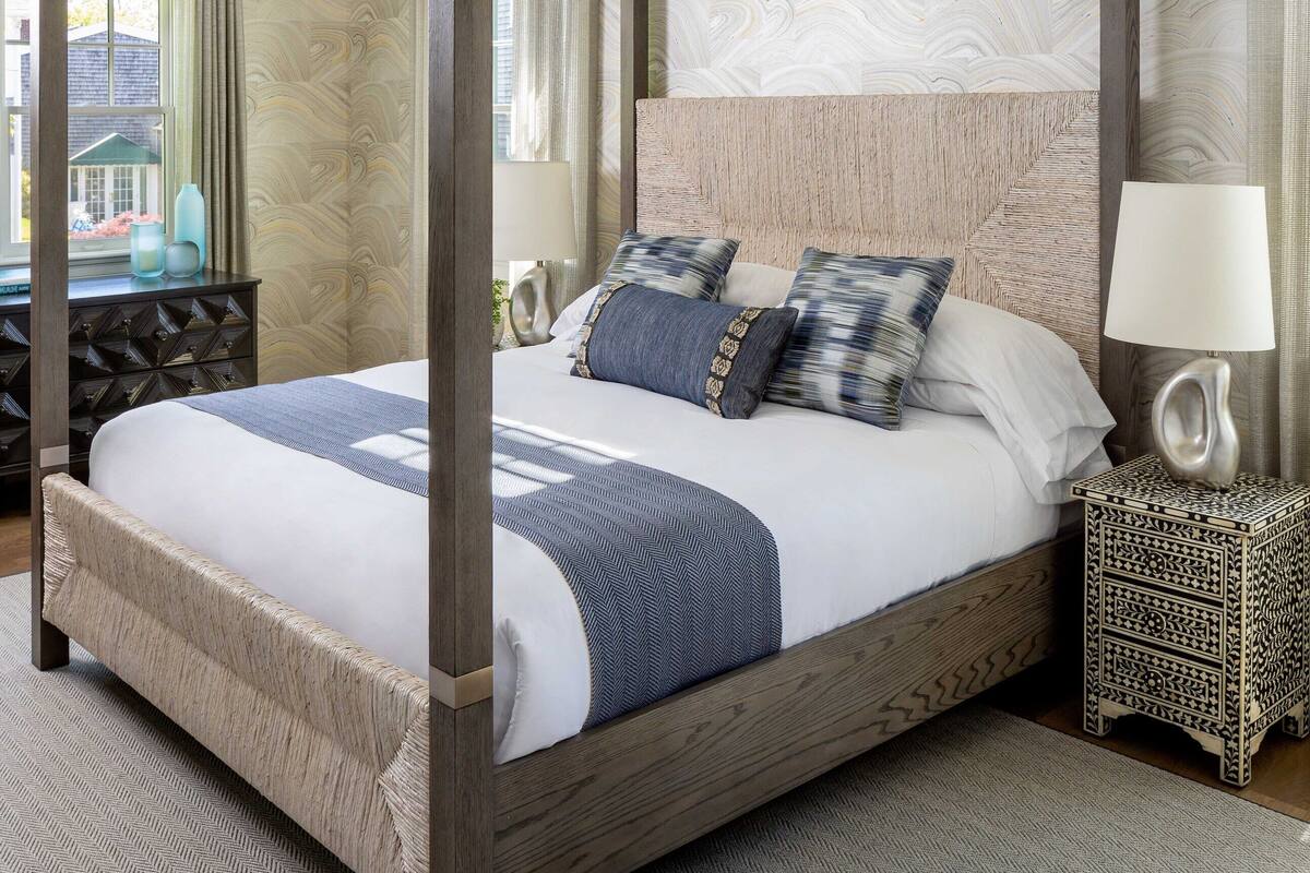 Una cama con dosel de madera y un cabecero texturizado, una mesita de noche con lámpara y vistas al exterior a través de una ventana.
