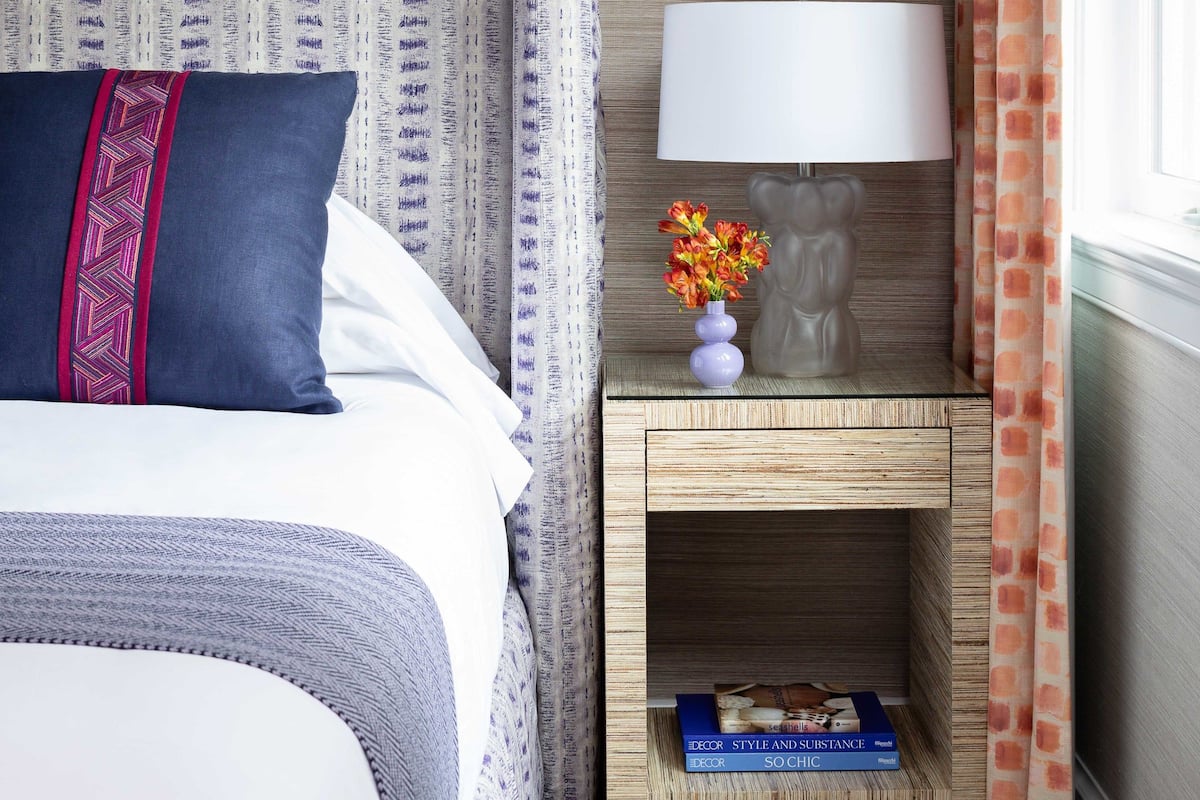 Una habitación de hotel impecable con una cama, una mesita de noche con lámpara, un jarrón con flores y un estante pequeño con libros.