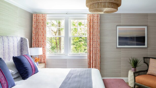 Une chambre d’hôtel avec un grand lit, une chaise, une fenêtre avec des rideaux, un tableau accroché au mur et un luminaire suspendu.