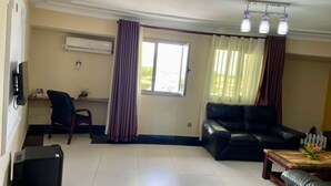 A living room with a black leather sofa, a wooden coffee table, and a TV on a stand. There is a window with curtains, a small table with a plant, and an air conditioning unit on the wall. - DK Hotel (Bonaberi)