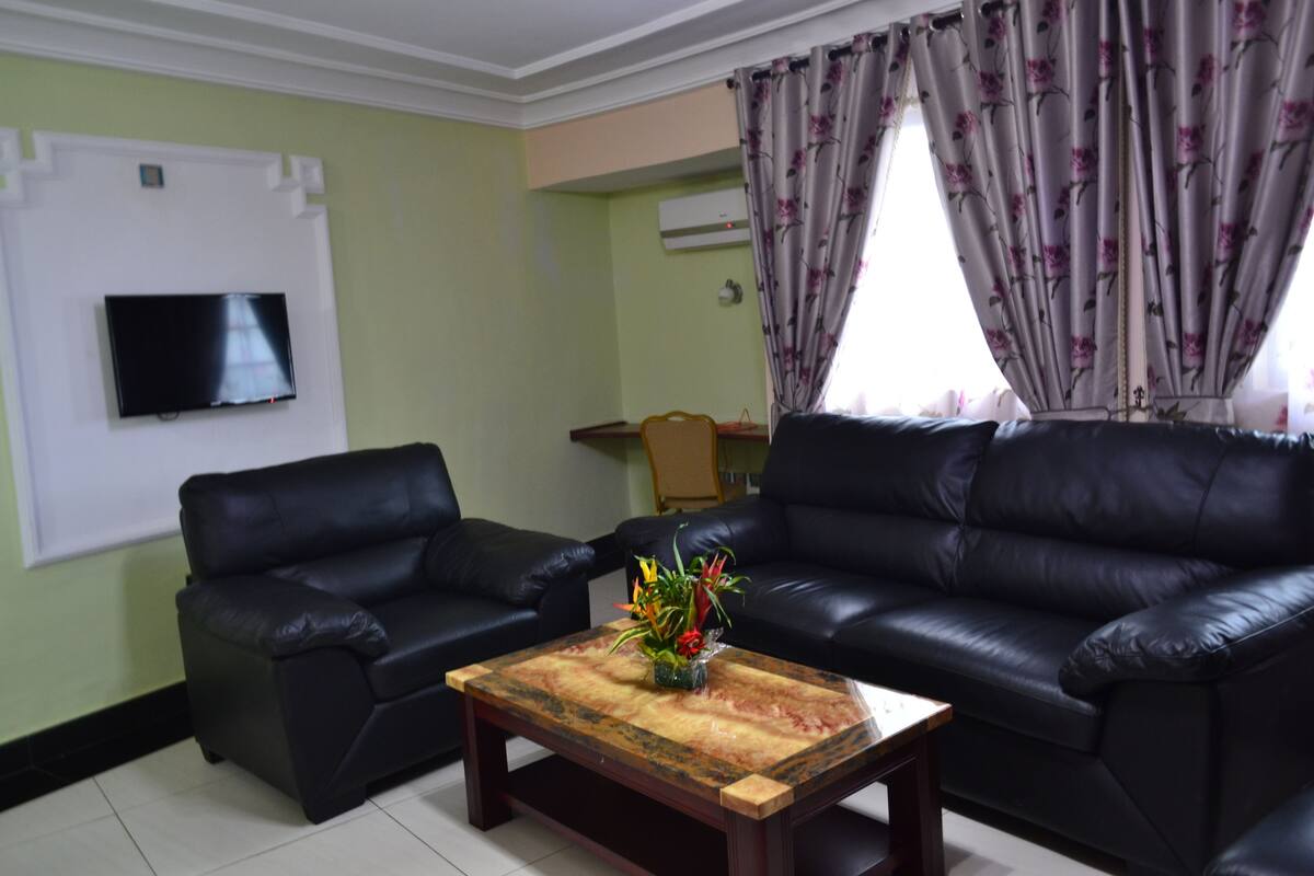 A living room with two black leather sofas, a wooden coffee table with a vase of flowers, a flat-screen TV on the wall, and a window with floral curtains.
