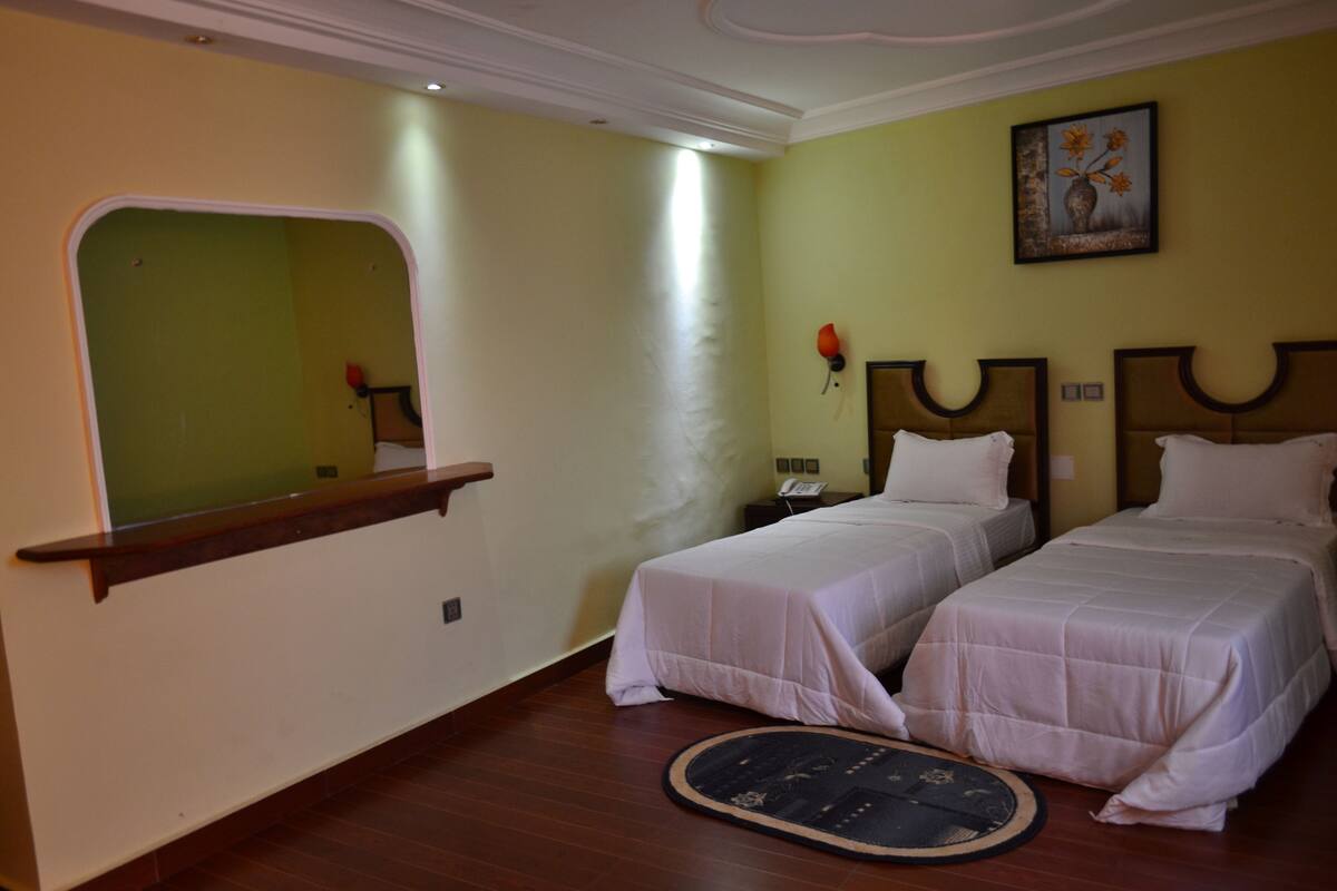 A hotel room with two single beds, a small desk, a mirror, and a painting on the wall.