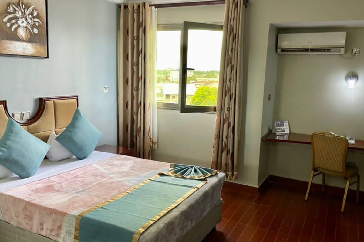 A hotel room with a bed, a desk, a chair, an air conditioner, and a window with curtains.