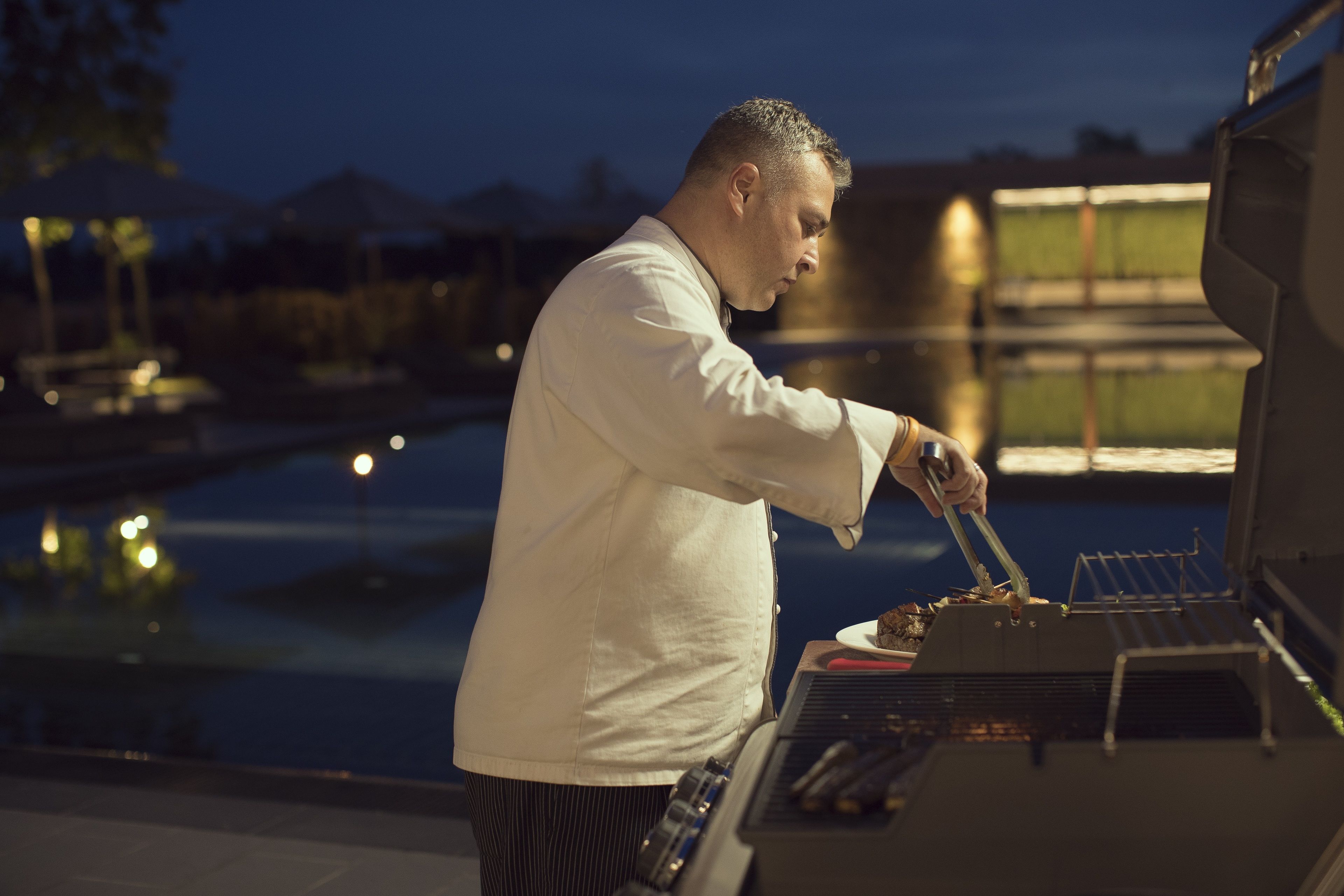 A person grilling outdoors at night with a pool in the background.
