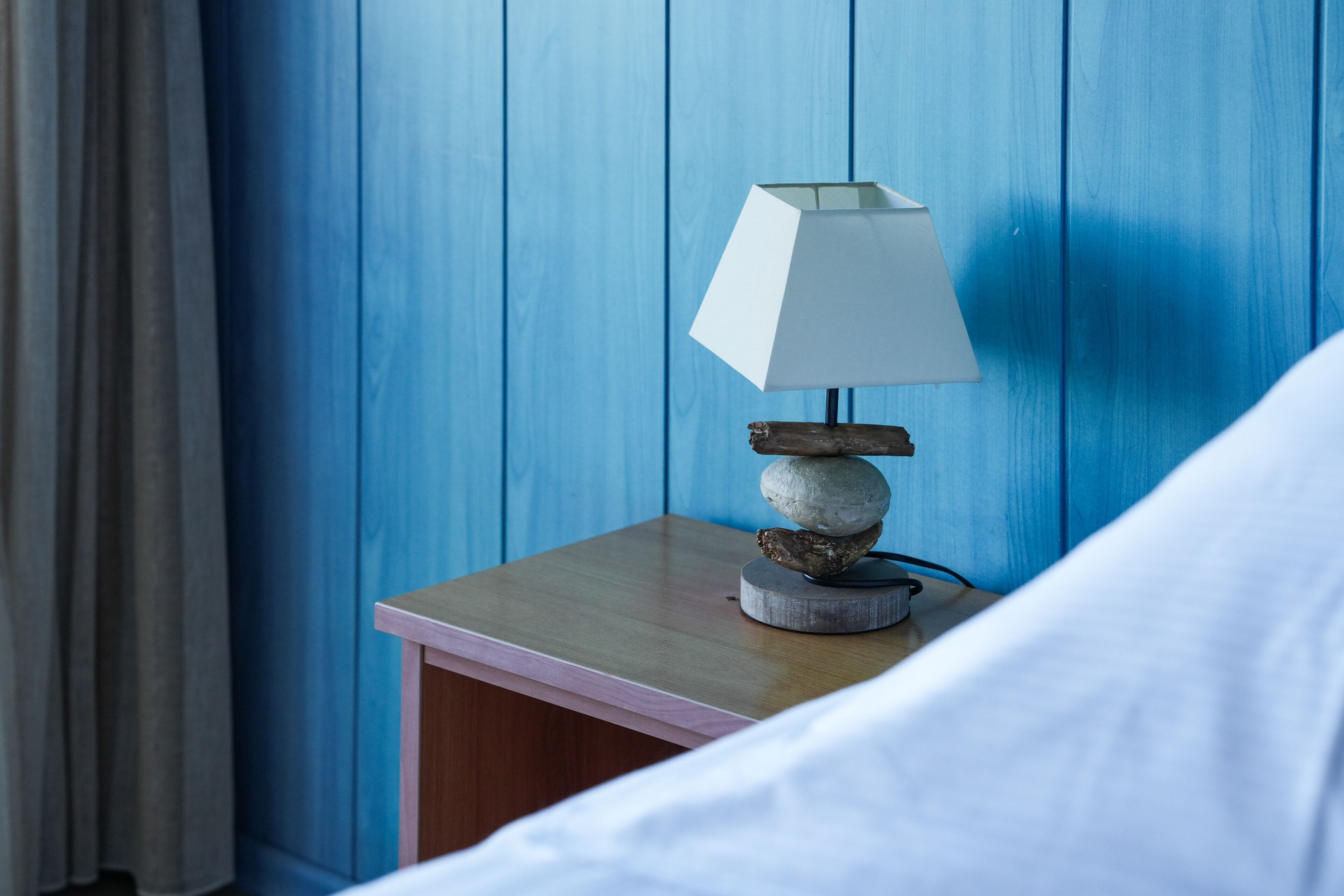 A neatly made bed with white linens, a bedside lamp with a white shade, and a wooden nightstand with a stack of stones.