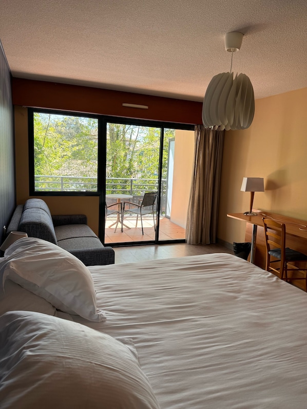 A hotel room with a large bed, a desk with a chair, a sofa, and a balcony with a table and chairs.