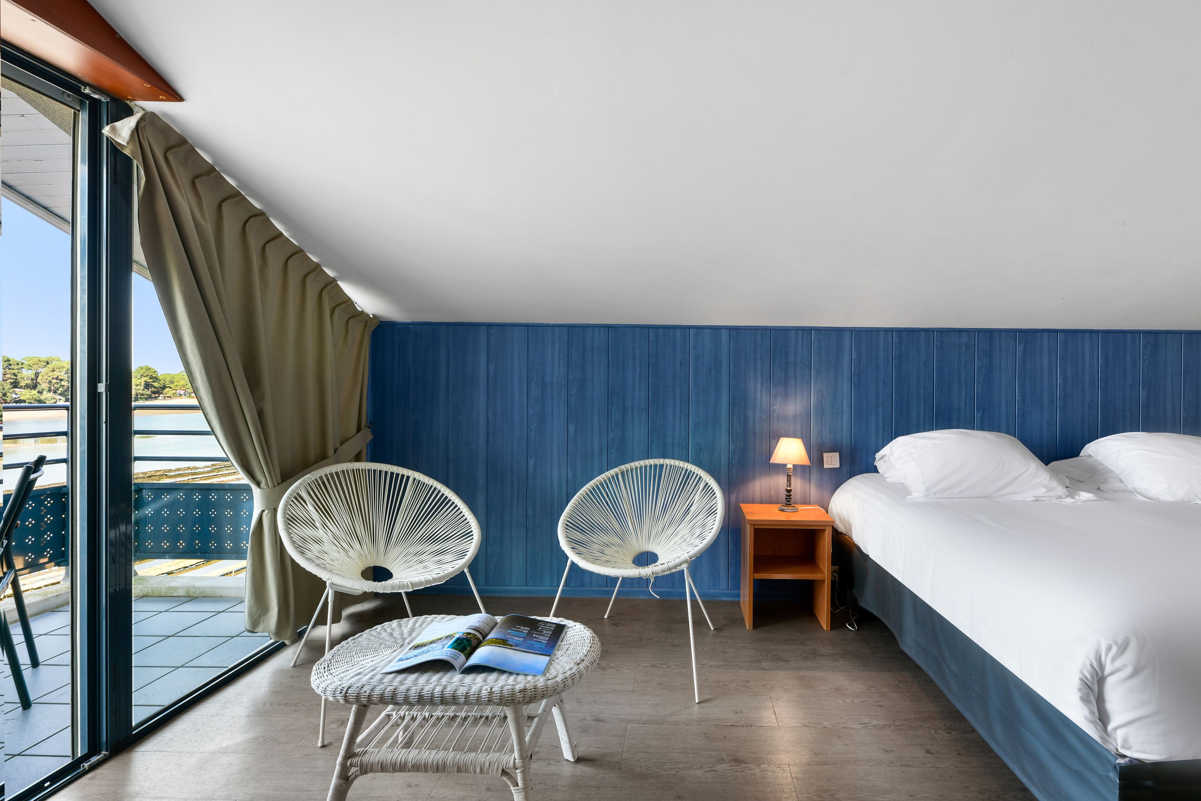 A hotel room with a bed, two wicker chairs, a small table with a lamp, and a view of the outdoors through a sliding door.
