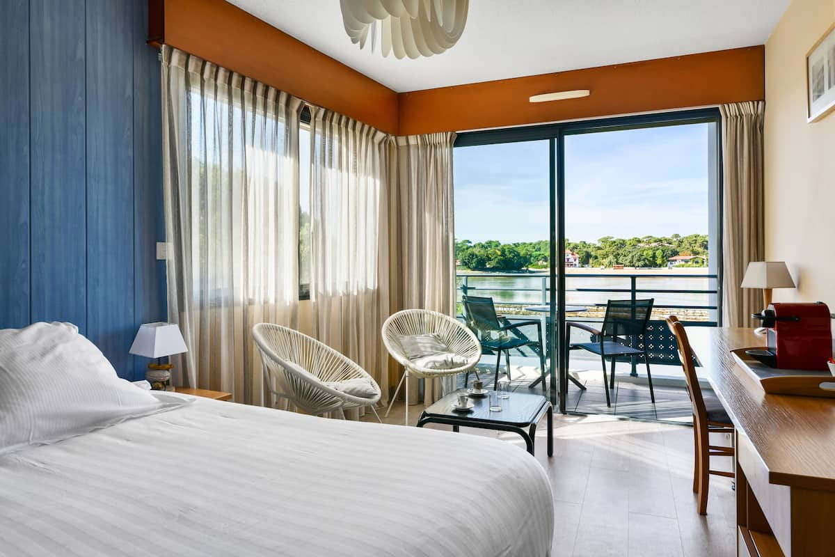 A hotel room with a large bed, two white wicker chairs, a small table with a lamp, and a view of a body of water through a sliding glass door.