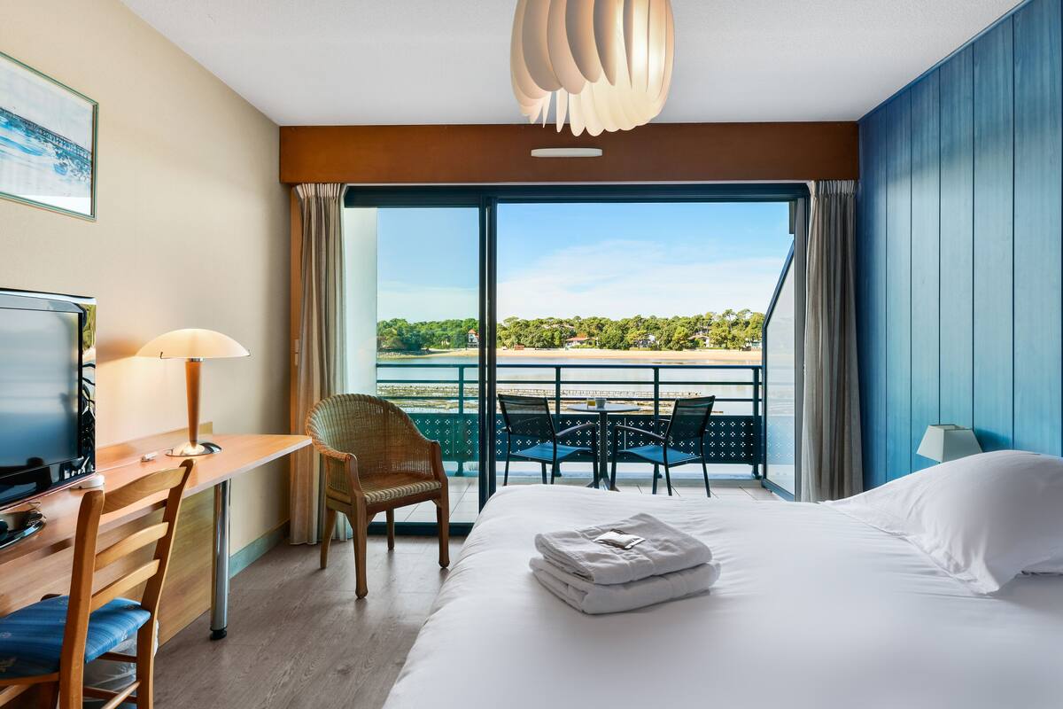 A hotel room with a bed, desk, chair, and a balcony with a view of the sea.