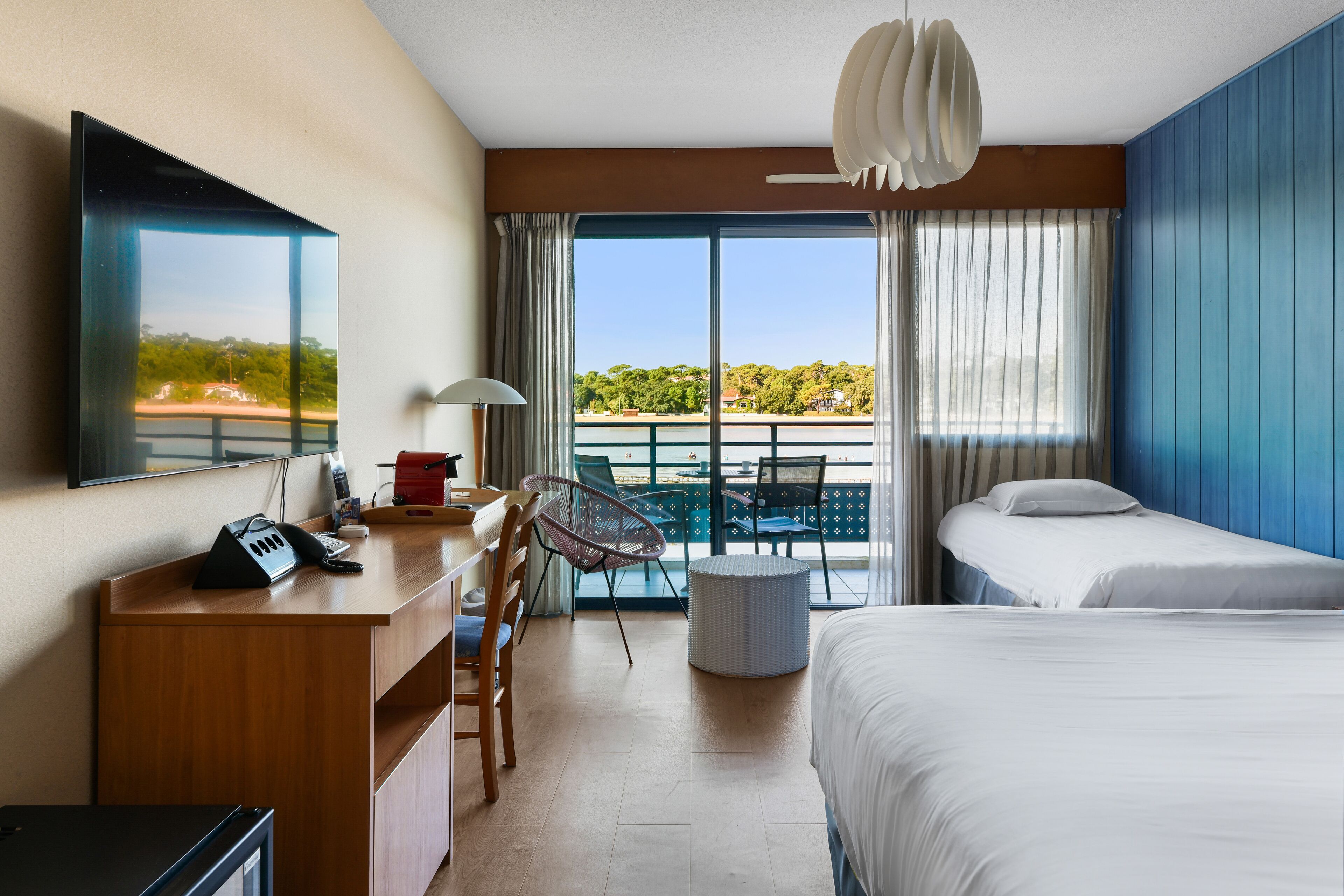 A hotel room with a desk, two beds, a balcony with a view, and a ceiling fan.