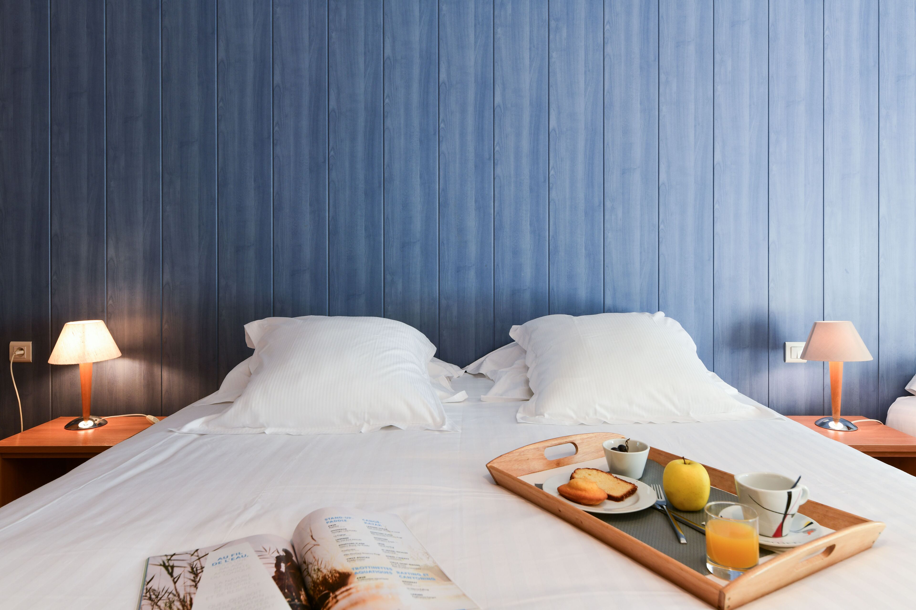 Ein Hotelzimmer mit einem Bett, weißen Kissen, einem Nachttisch aus Holz mit einer Lampe, einer Ablage mit Frühstücksgegenständen und einer Zeitschrift auf dem Bett.
