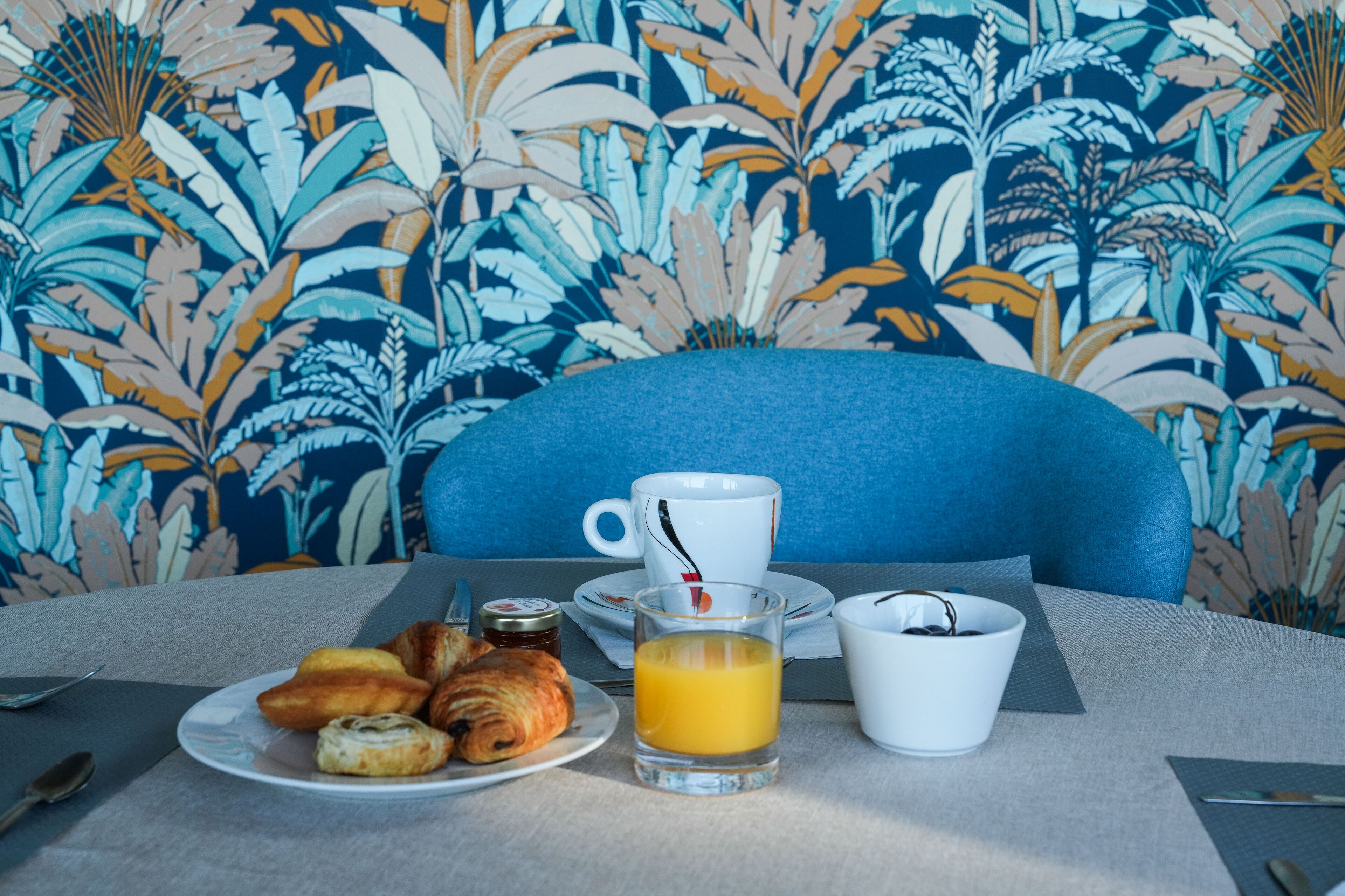 A breakfast setup with croissants, orange juice, and a cup on a table with a blue chair and a patterned wallpaper background.