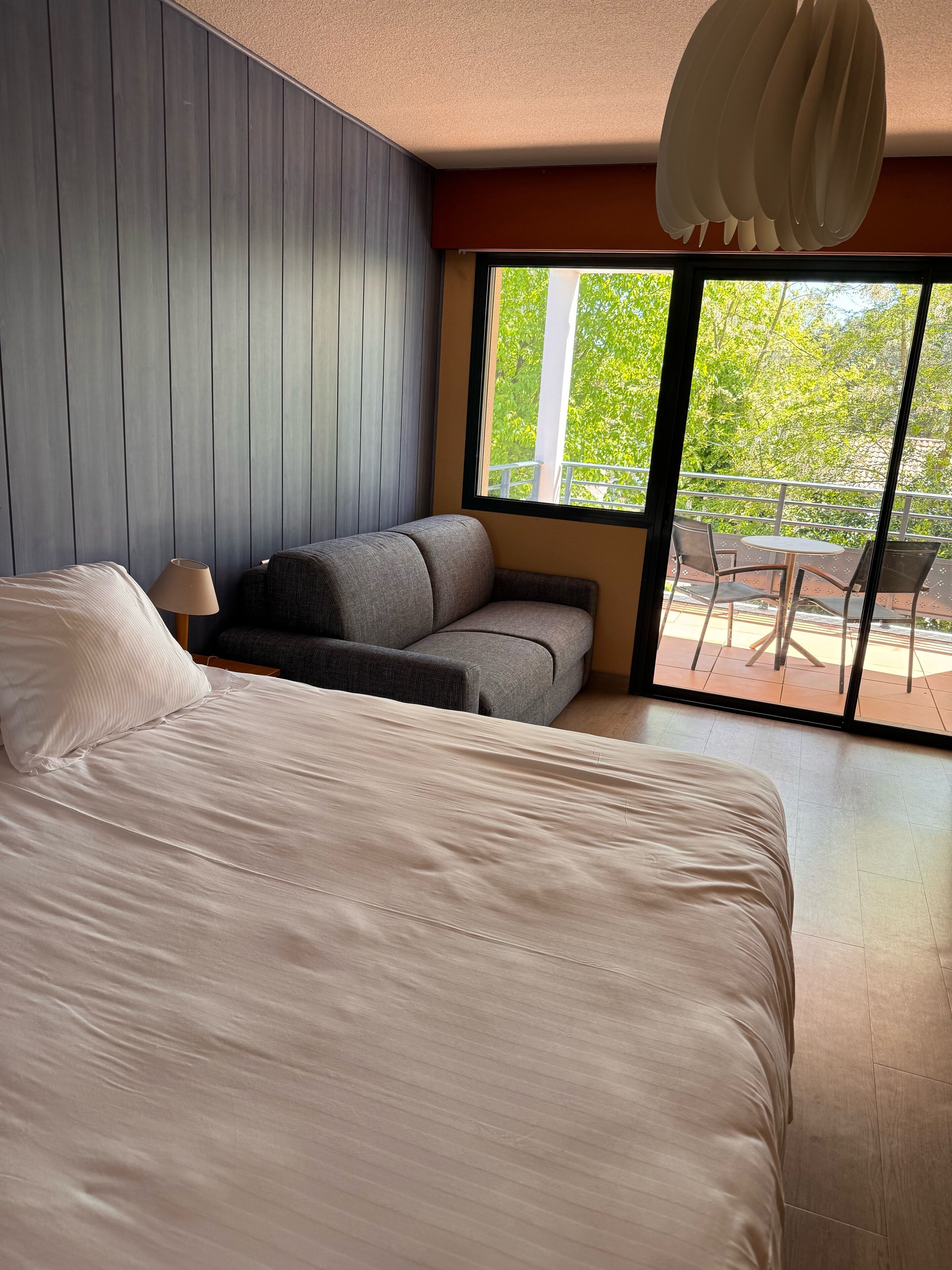 A modern hotel room with a large bed, a sofa, and a balcony with outdoor seating.