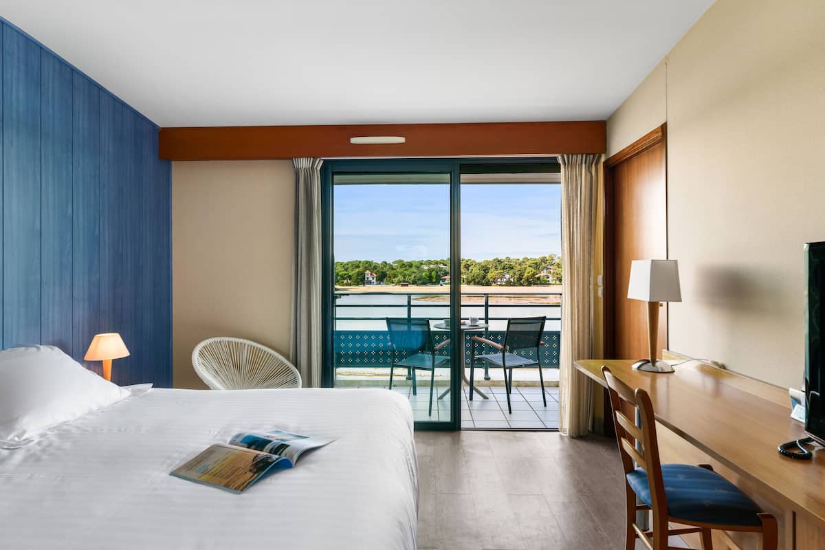 A hotel room with a bed, a desk, a chair, a TV, and a balcony with a view of the sea.