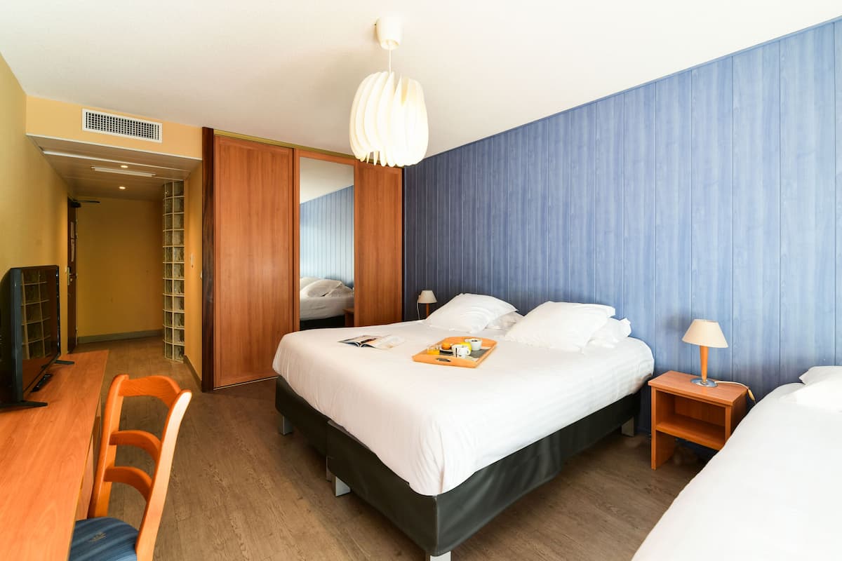 A hotel room with a large bed, a desk, a chair, a nightstand with a lamp, and a wardrobe.