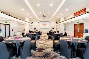 Egy konferenciaterem, melyben kerek asztalok, székek és egy projektorvászon található, amelyen a „HARRIS Seminyak Bali” felirat látható.