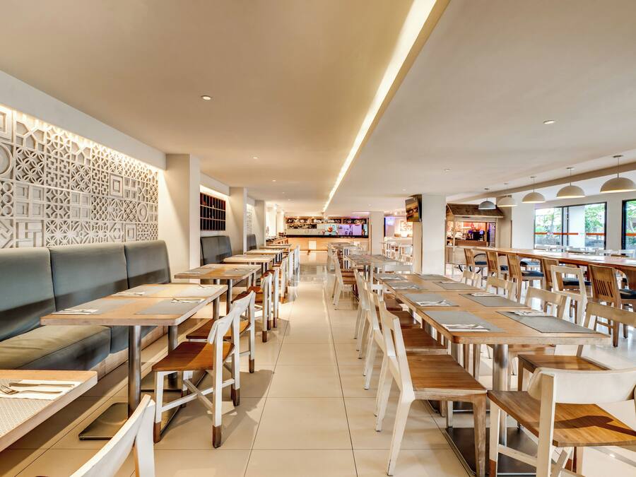 Interior moderno de restaurante con mesas y sillas altas, zona de estar con banco acolchado y pared decorativa con patrones geométricos.