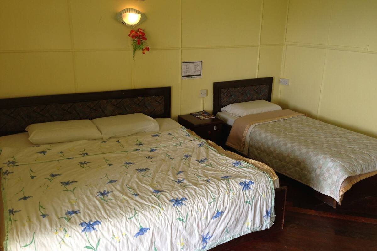Ein Zimmer mit zwei Betten, jeweils mit einem Holz-Kopfteil und einer blumigen Tagesdecke. Zwischen den Betten befindet sich ein Nachttisch mit einer Lampe.