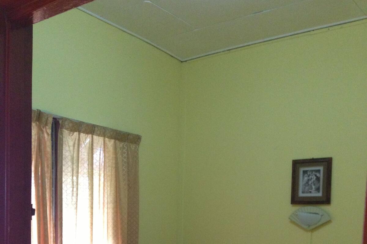 Ein kleines, schlichtes Zimmer mit einem Bett, einem Nachttisch und einem Fenster mit Vorhängen.