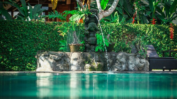 A stone fountain with water spouting from it, surrounded by lush greenery and a pool. - Nick's Pension (Ubud)