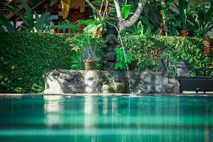 A stone fountain with water spouting from it, surrounded by lush greenery and a pool.