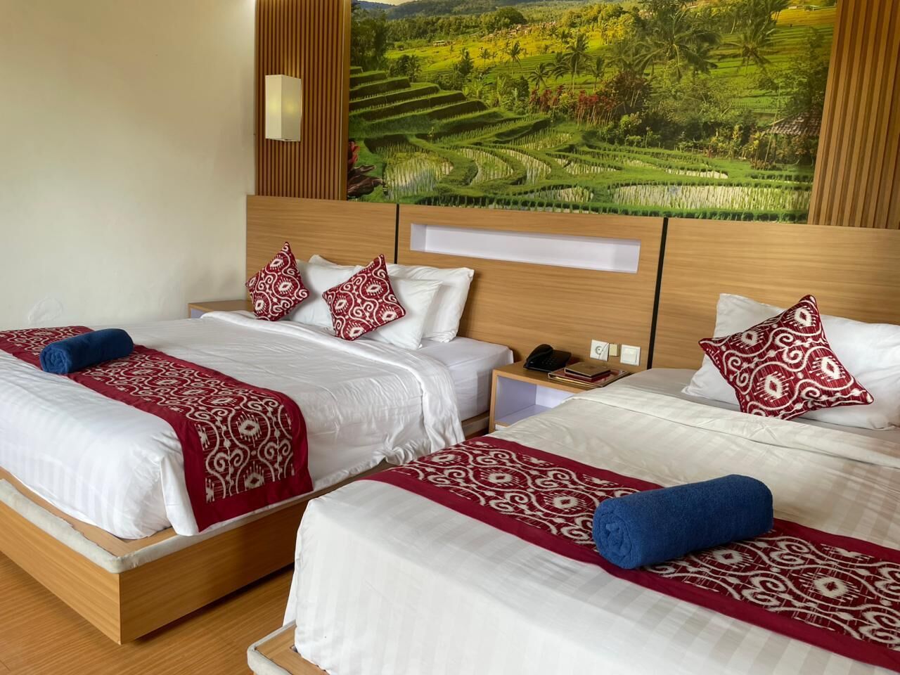 A hotel room with three beds, each with a patterned bedspread and pillows, a wooden headboard, and a large landscape painting above the beds.