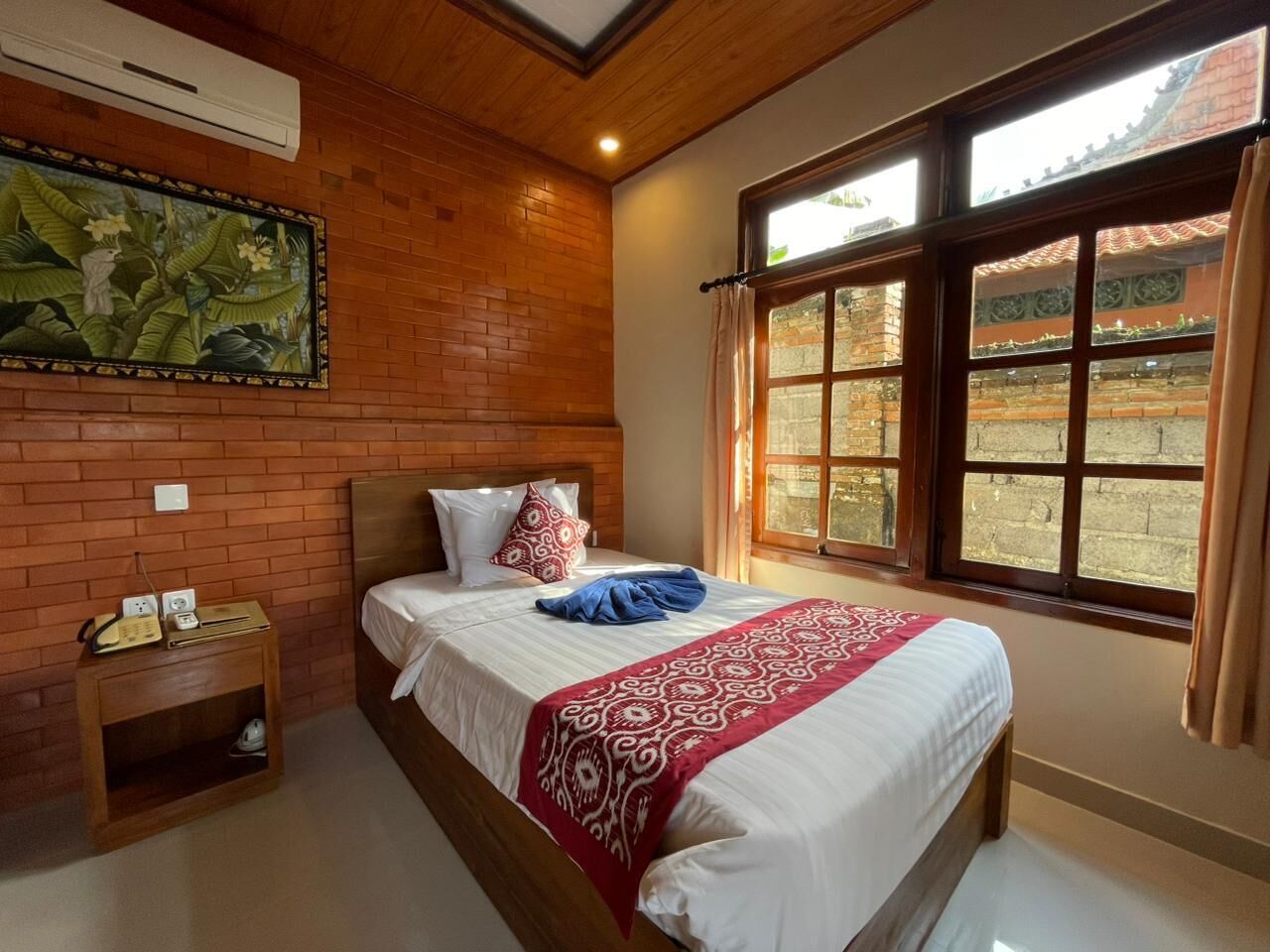 A hotel room with a brick wall, a bed with white bedding and a red patterned throw, a nightstand with a telephone, and a window with a view of a building.