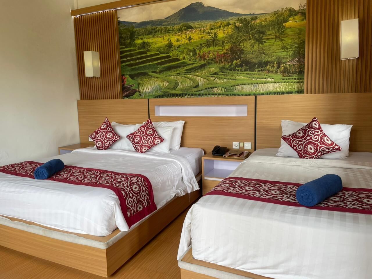 A hotel room with two beds, each with a wooden headboard, white bedding, and patterned red and blue throw pillows. A large landscape painting is on the wall above the beds.