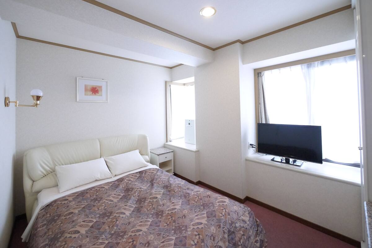 A hotel room with a bed, a television on the wall, a window, and a bedside table.