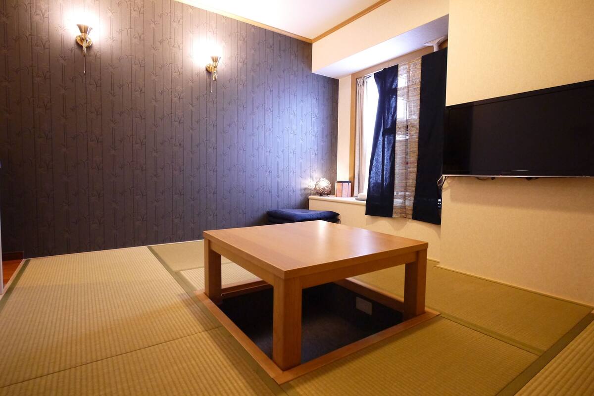 A room with tatami mat flooring, a wooden table, a wall-mounted TV, and a window with curtains.