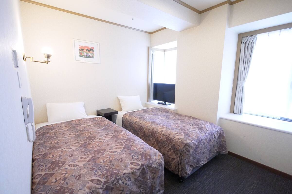A hotel room with two beds, a TV, a telephone, and a window with curtains.