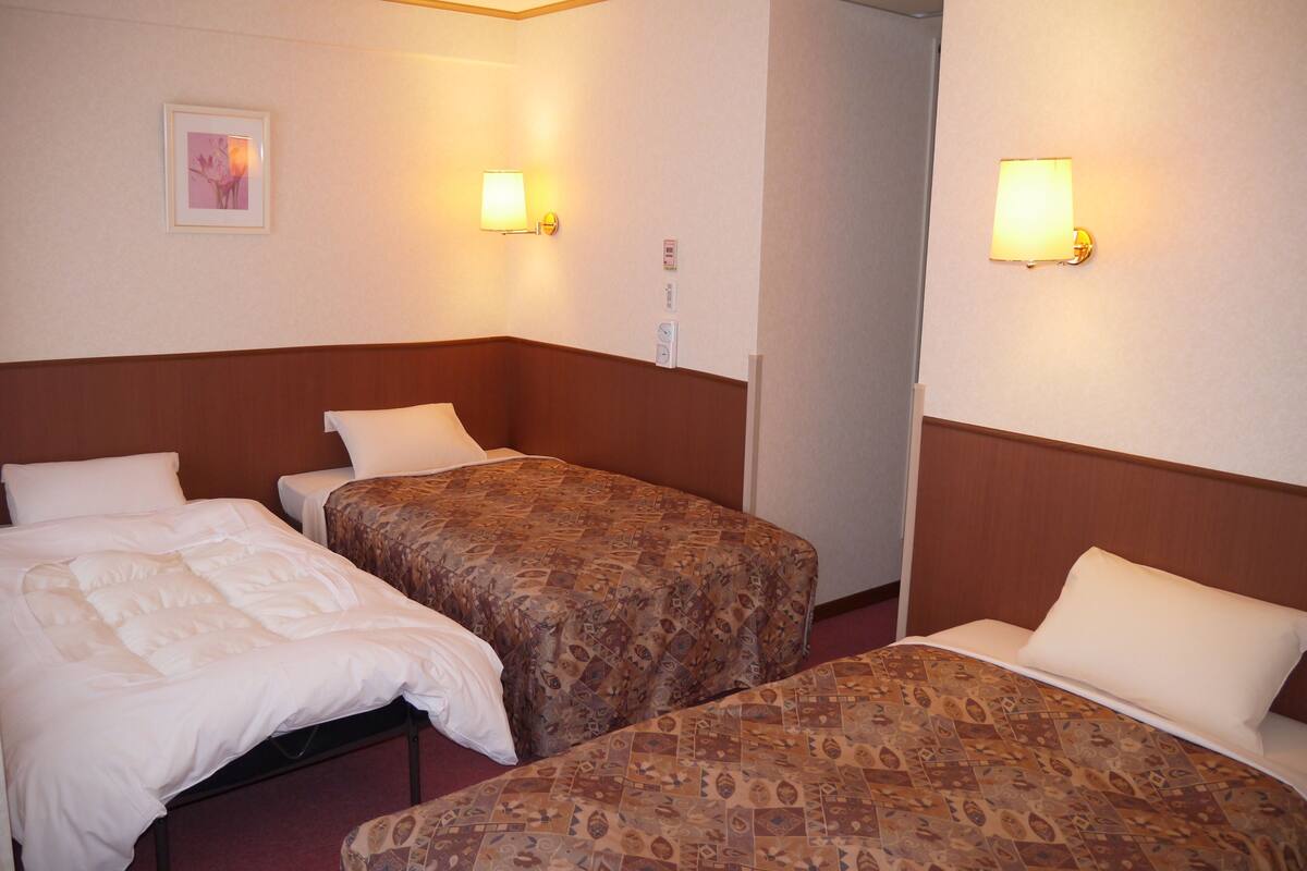 A hotel room with two single beds, each with a patterned bedspread and white pillows. There are two wall-mounted lamps providing light.