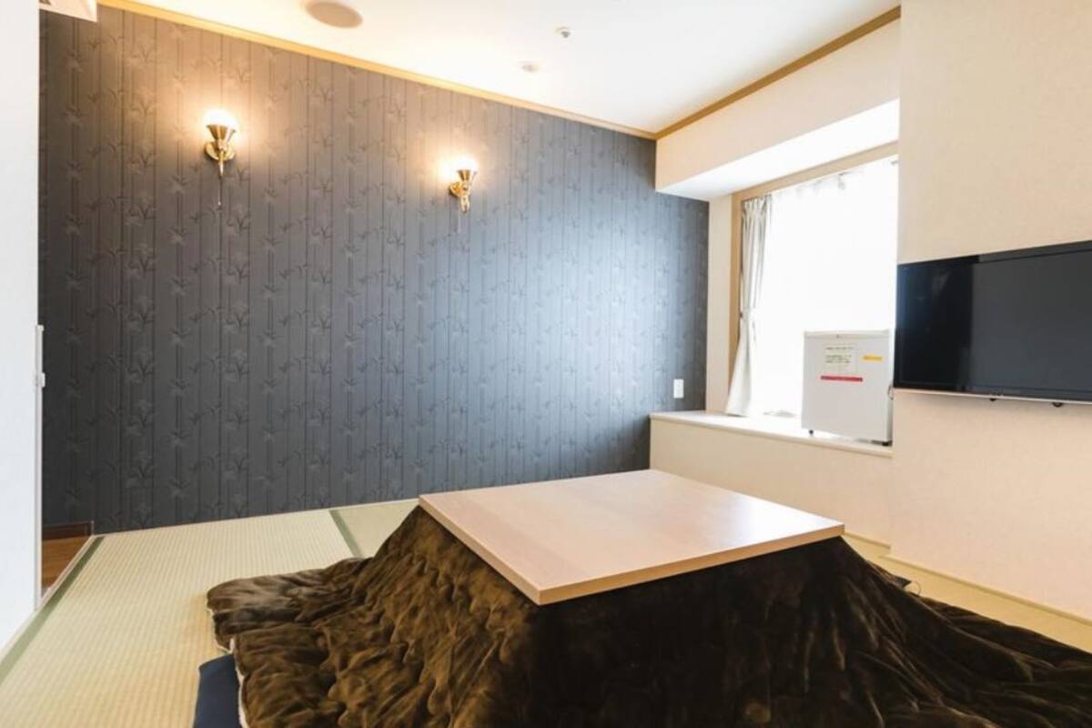 A room with a wooden floor, a flat-screen TV mounted on the wall, a window with curtains, and a small table.
