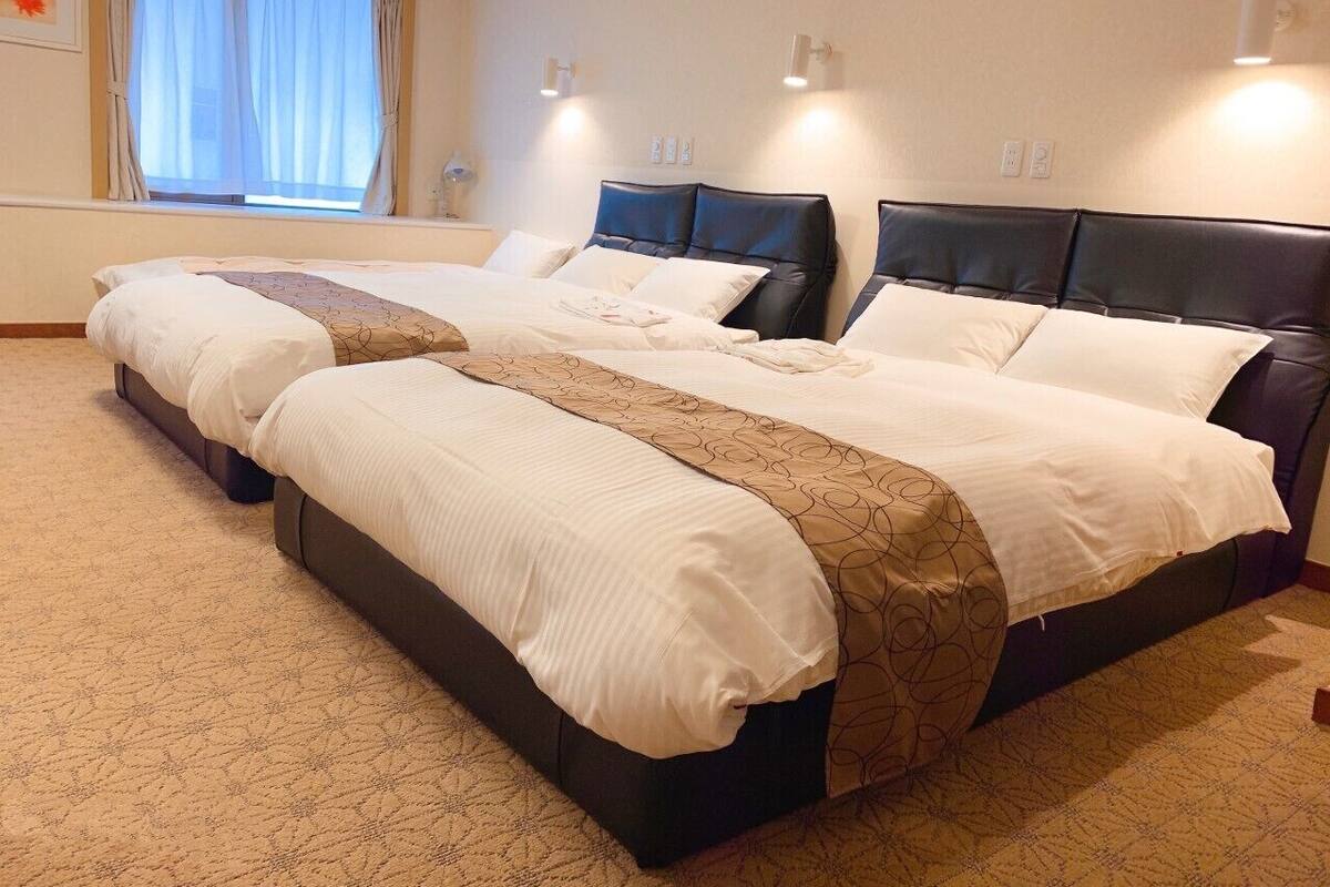 A hotel room with two beds, a large window with curtains, and wall-mounted lighting.