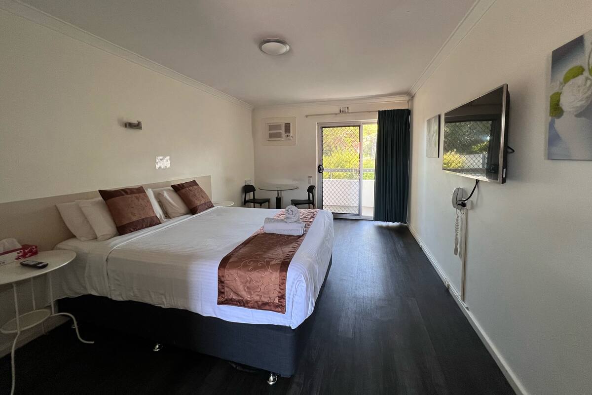 A hotel room with a large bed, a desk, a chair, a TV, and a balcony with a view.