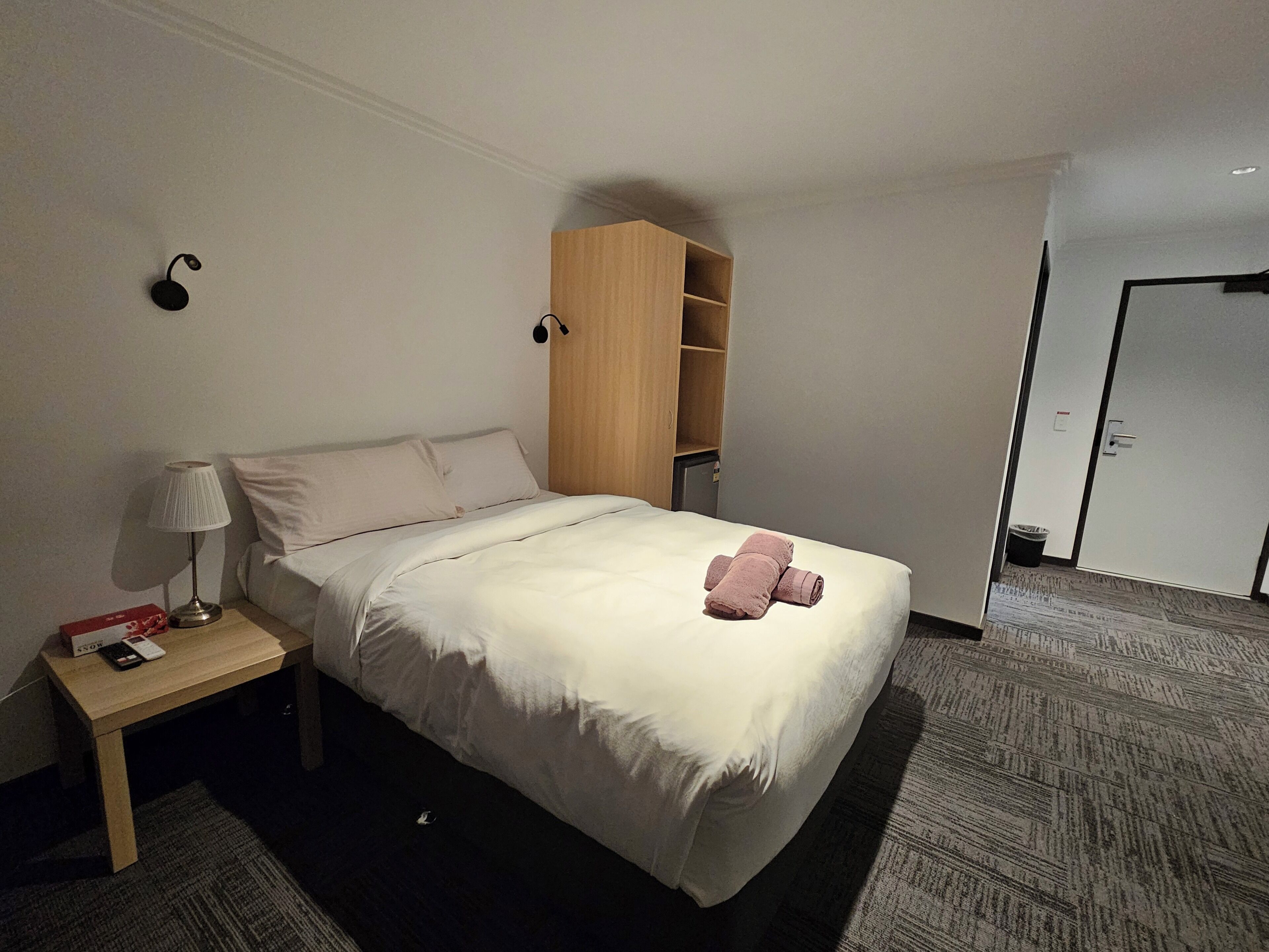 A hotel room with a bed, a nightstand with a lamp, a wooden wardrobe, and a door.