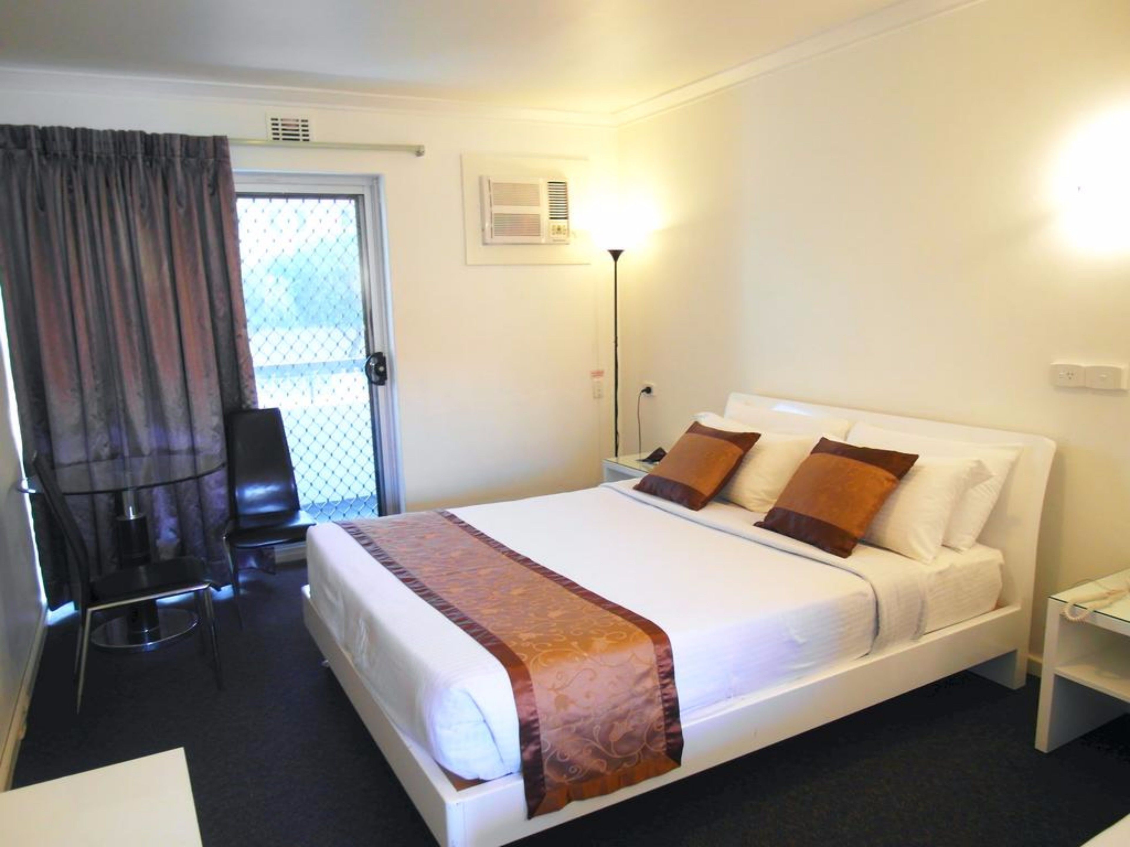 Una habitación de hotel con cama, mesita de noche, lámpara, silla y ventana con cortinas.