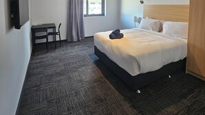 A hotel room with a bed, a desk, a chair, a window with curtains, and a wall-mounted air conditioner.