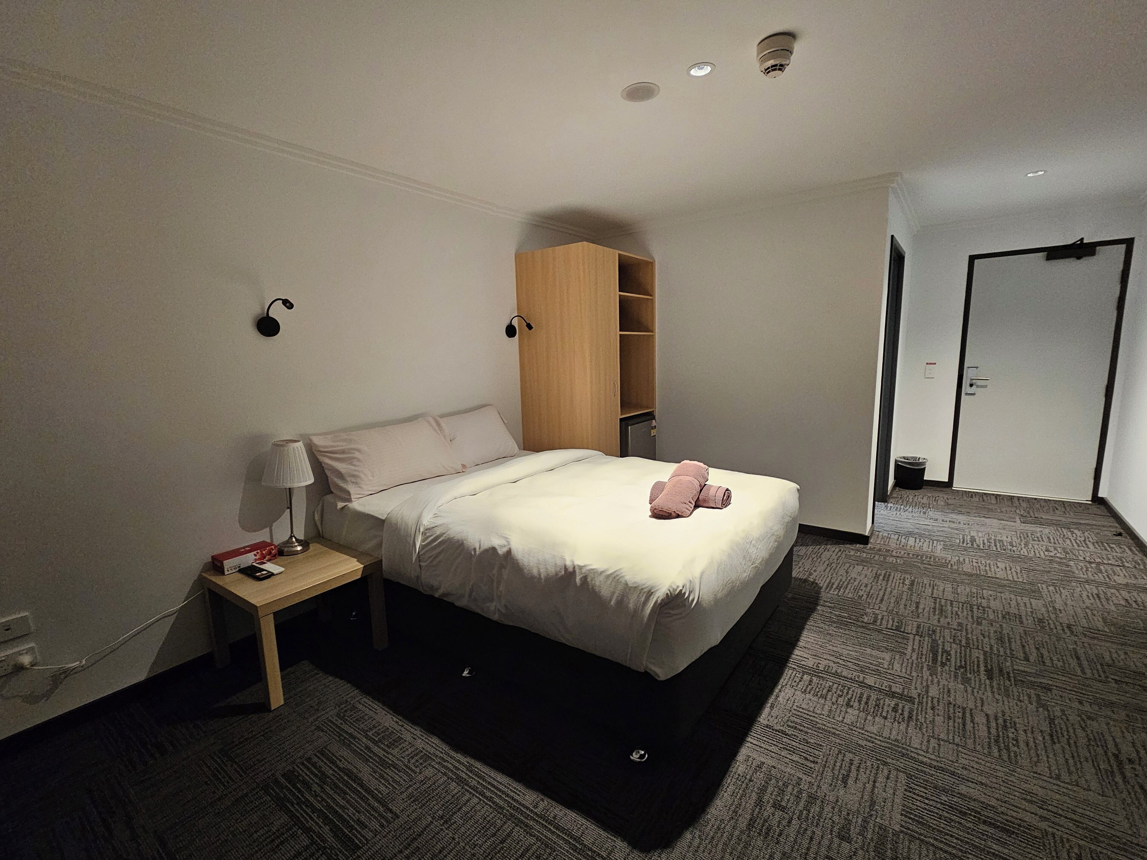 Una camera d'albergo ordinata con un letto, un comodino, una lampada e un armadio.
