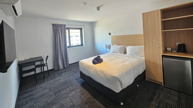 A hotel room with a bed, a desk, a chair, a wardrobe, a small kitchenette, and a window with curtains.