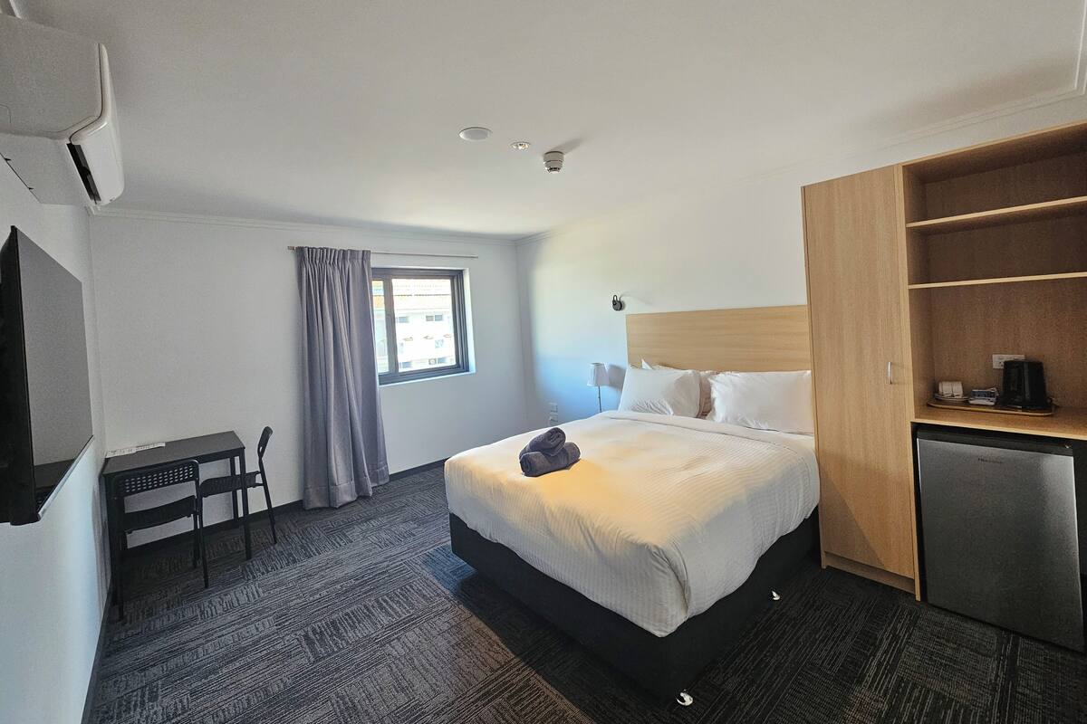 A hotel room with a bed, a desk, a chair, a wardrobe, a small kitchenette, and a window with curtains.