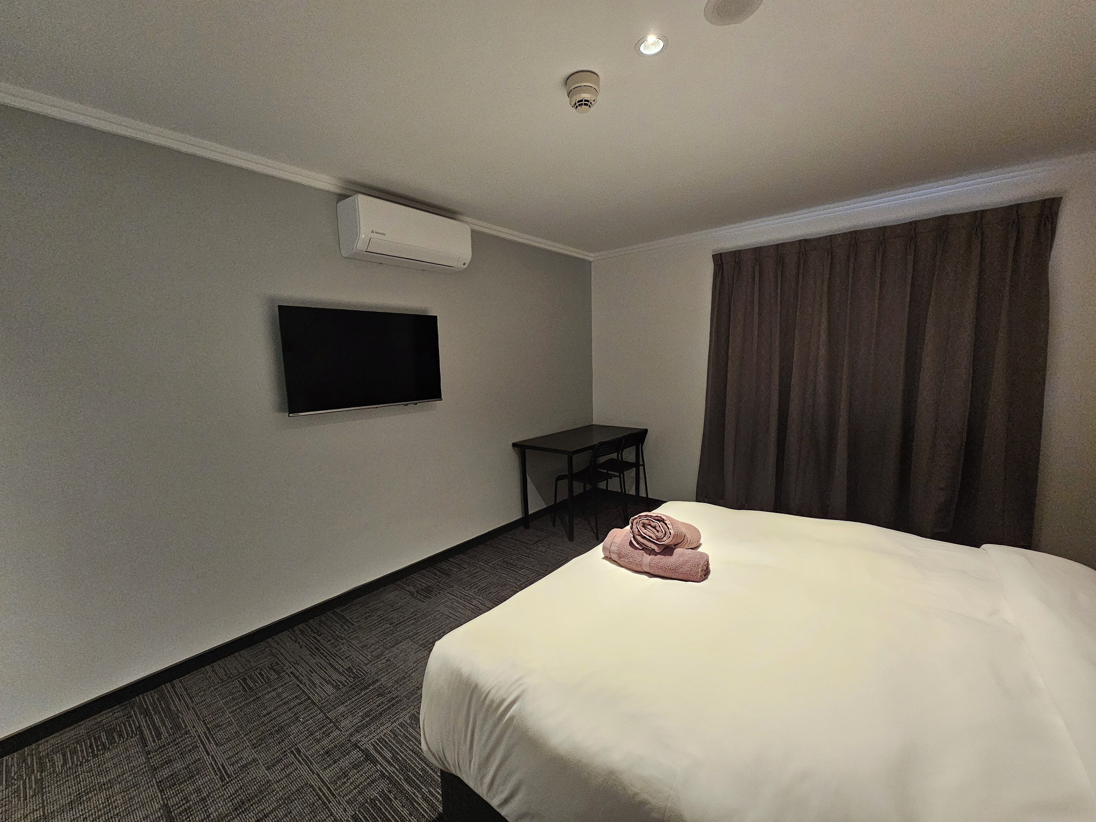 A hotel room with a bed, a flat-screen TV mounted on the wall, a small table, and a chair.
