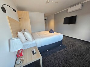 A hotel room with a bed, a bedside table with a lamp, a TV, and a wall-mounted air conditioner.