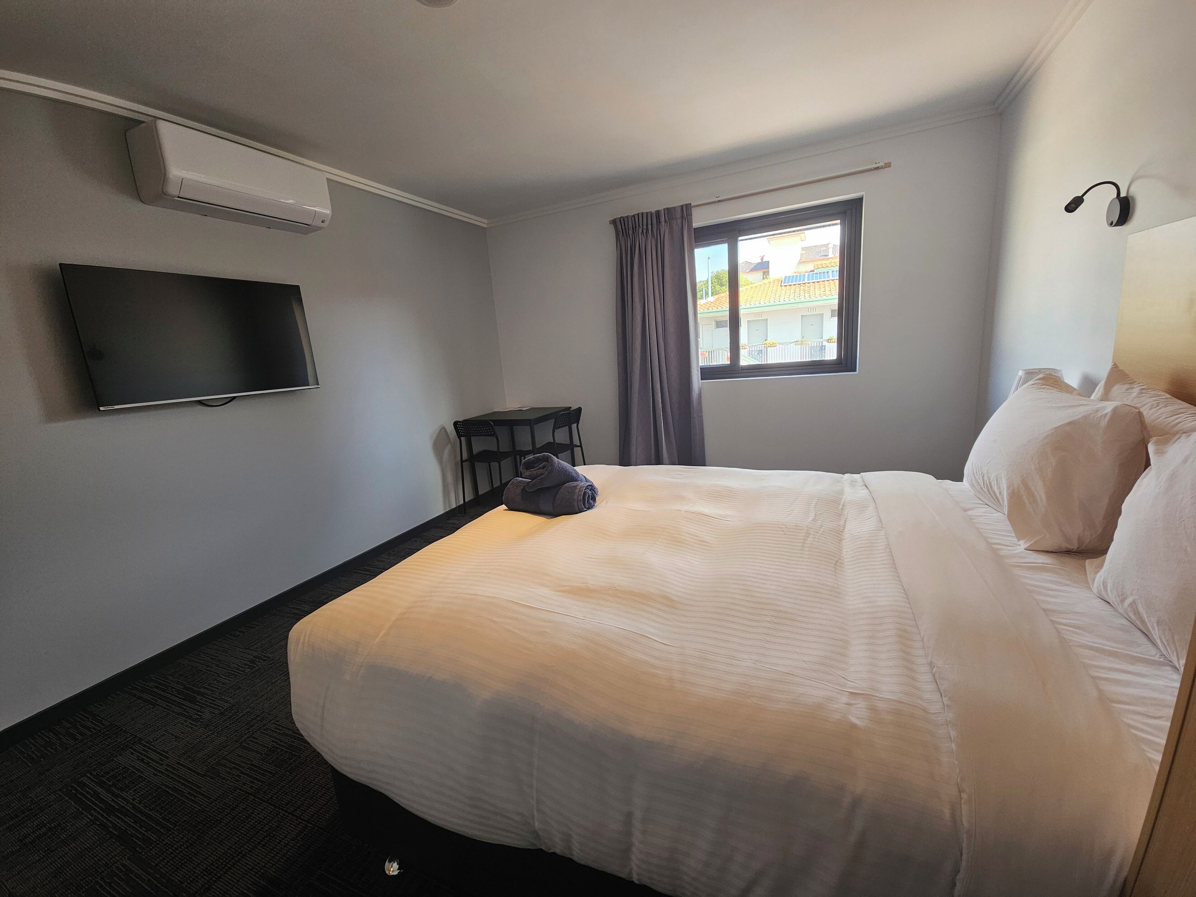 Habitación de hotel con una cama grande, un televisor de pantalla plana montado en la pared, una mesita y una ventana con cortinas.