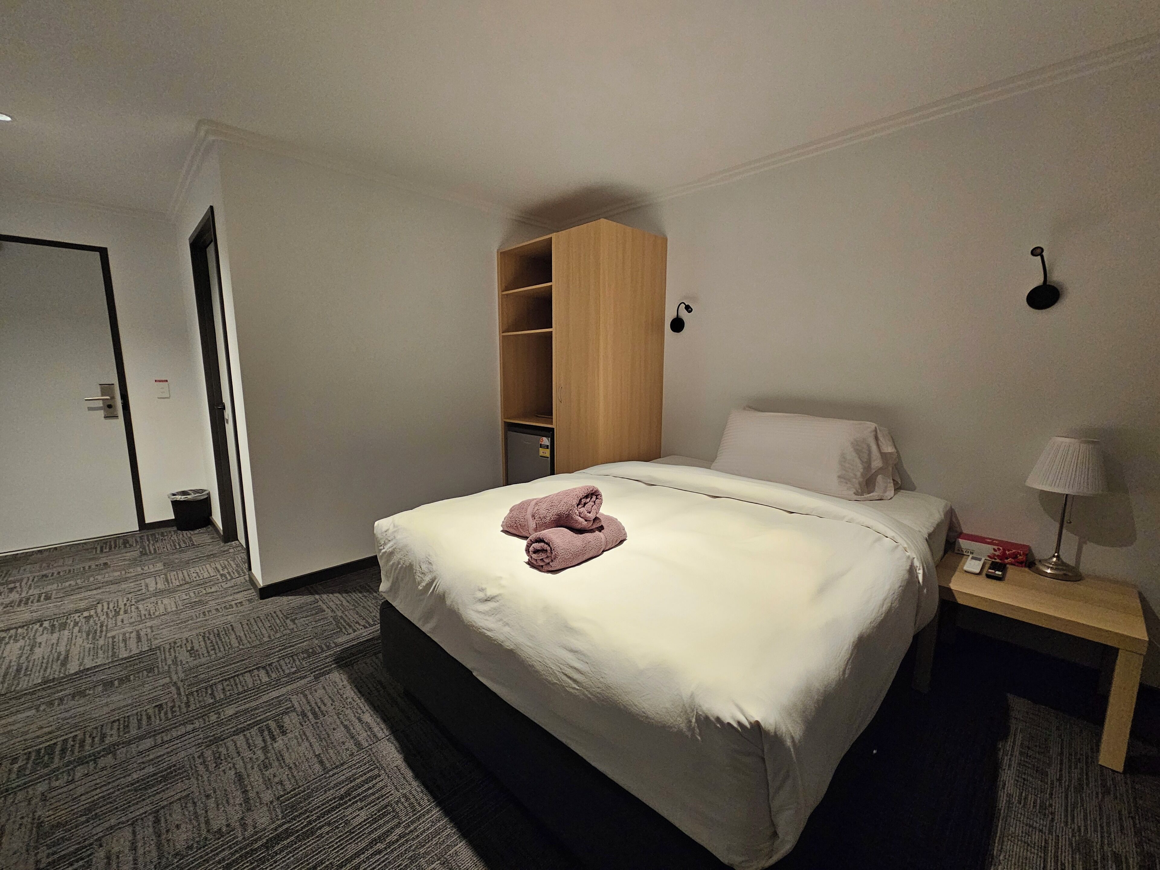 A hotel room with a bed, a bedside table with a lamp, a wardrobe, and a door.