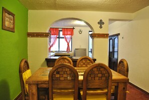 A dining area with a wooden table and chairs, a window with curtains, and a cross on the wall. - Casa Geranios Tequisquiapan (Tequisquiapan)