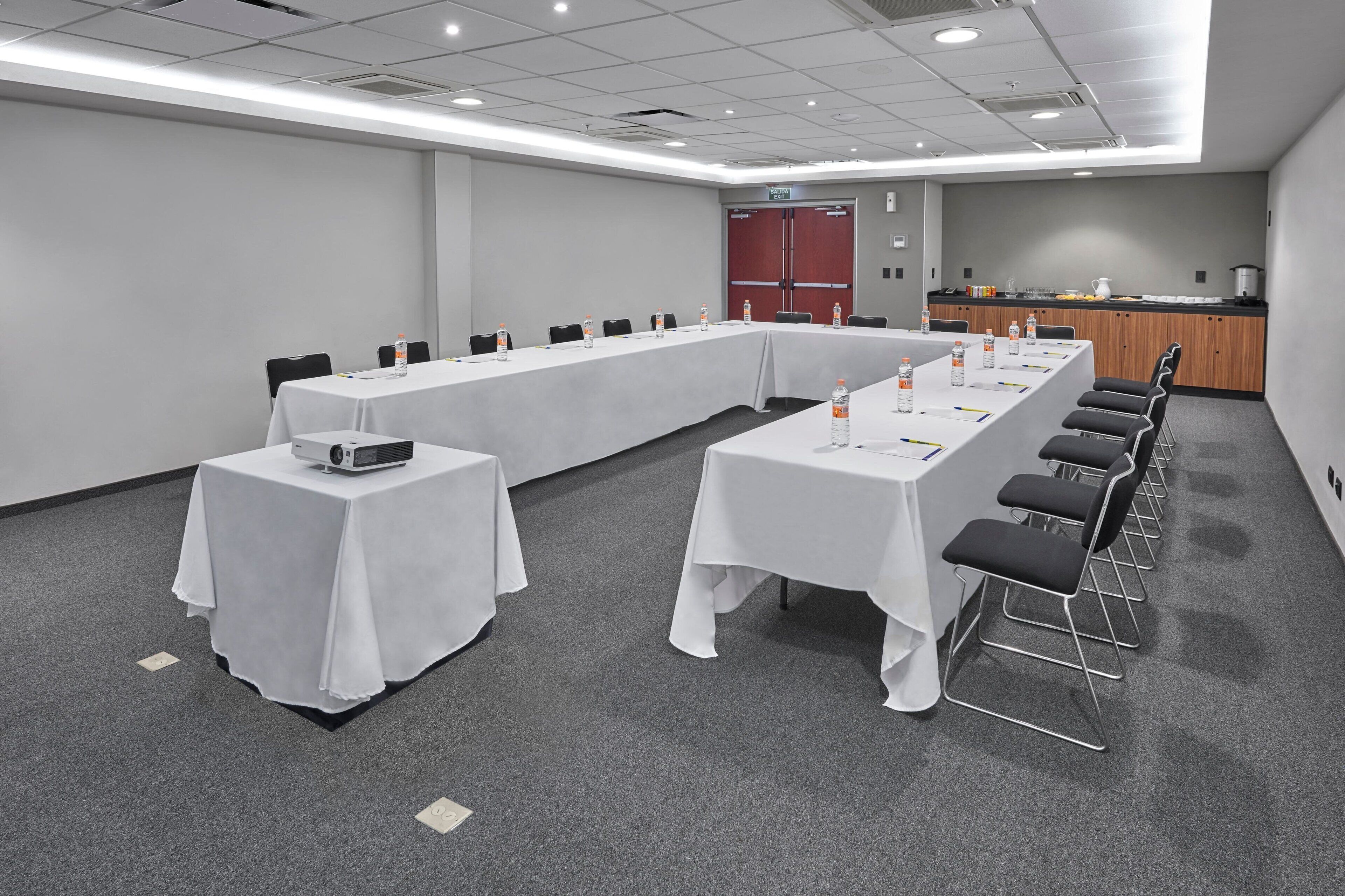 Un salón de conferencias con una mesa larga, sillas, un proyector y una zona de kitchenette.