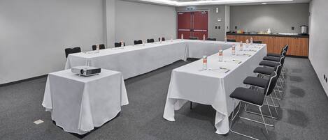 Un salón de conferencias con una mesa larga, sillas, un proyector y una zona de kitchenette.