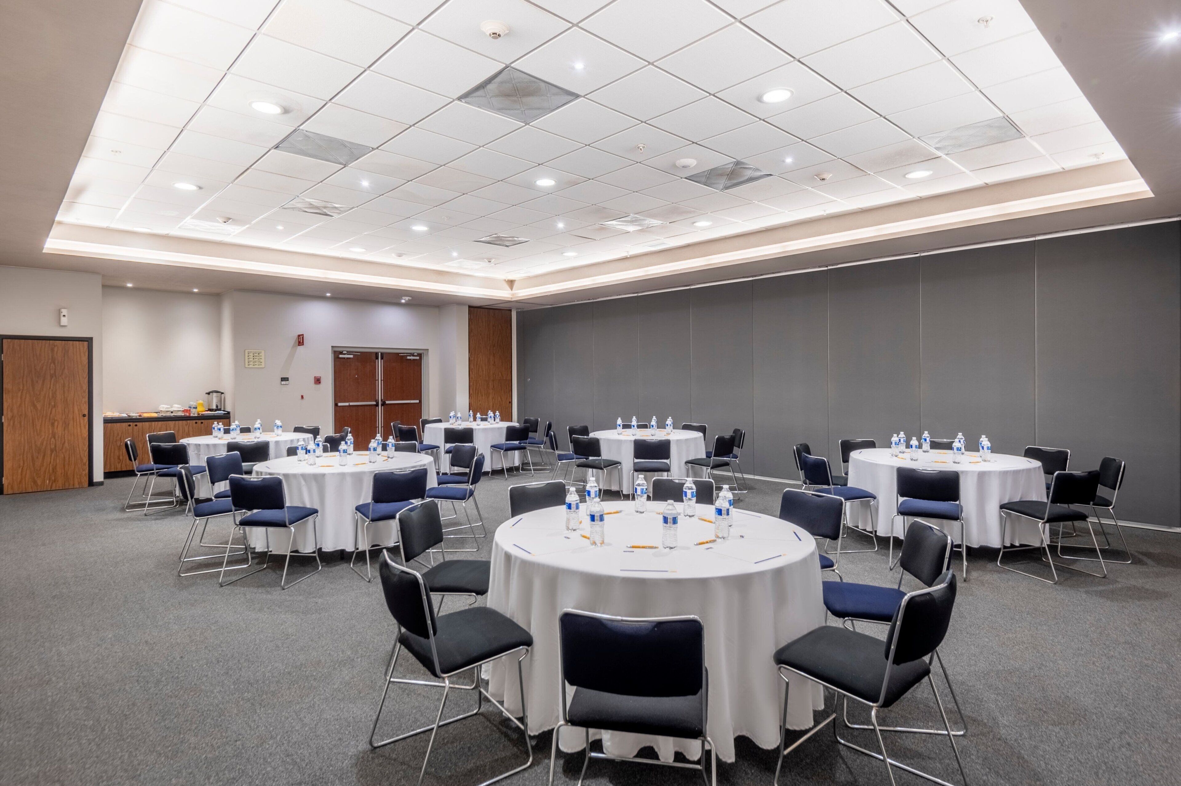 A conference room with multiple round tables, each surrounded by chairs, and equipped with water bottles and notepads.