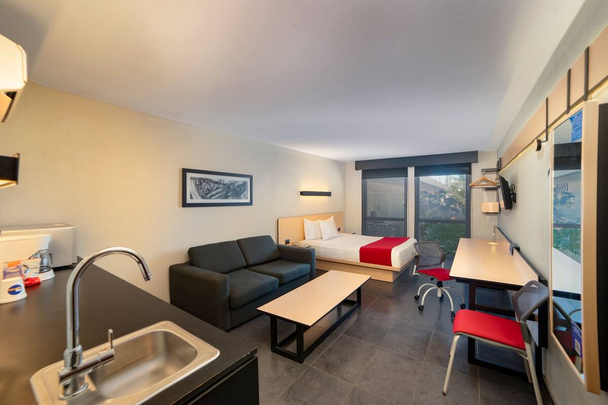 A modern hotel room with a kitchenette, a sofa, a bed with a red blanket, a desk with a chair, and large windows offering a city view.